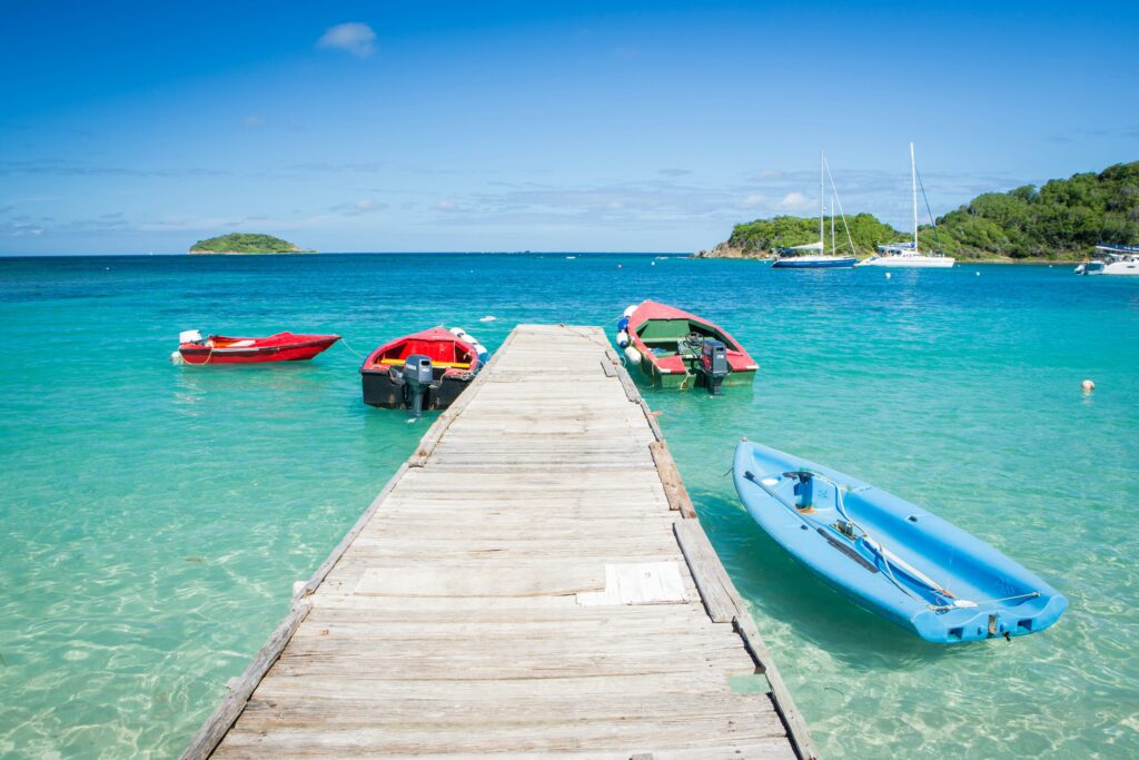 A foto destaca um píer rústico em Bequia, ilha de São Vicente e Granadinas, ladeado por lanchas vermelhas, caiaque azul e veleiros ancorados em mar turquesa—cenário perfeito para passeios de barco, mergulho e turismo náutico no Caribe. Palavras-chave: Bequia, São Vicente e Granadinas, píer de madeira, águas cristalinas, passeio de barco no Caribe.