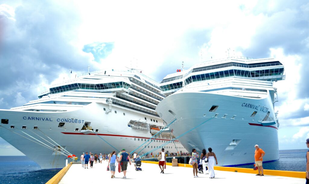 Vista frontal de dois navios de cruzeiro brancos da Carnival, o Carnival Conquest e o Carnival Valor, amarrados a um píer. O céu está parcialmente nublado e há pessoas caminhando na doca.