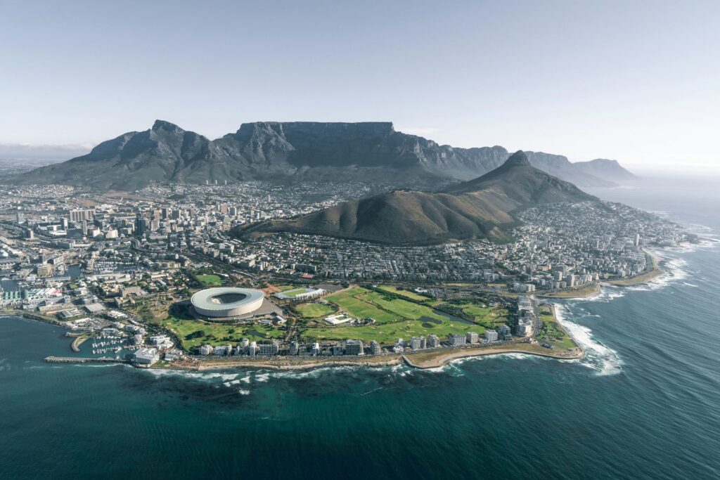 A imagem captura uma vista aérea impressionante da Cidade do Cabo, África do Sul, destacando a icônica Table Mountain, o Estádio Cape Town e a fusão harmoniosa entre a vibrante paisagem urbana e a beleza natural costeira.