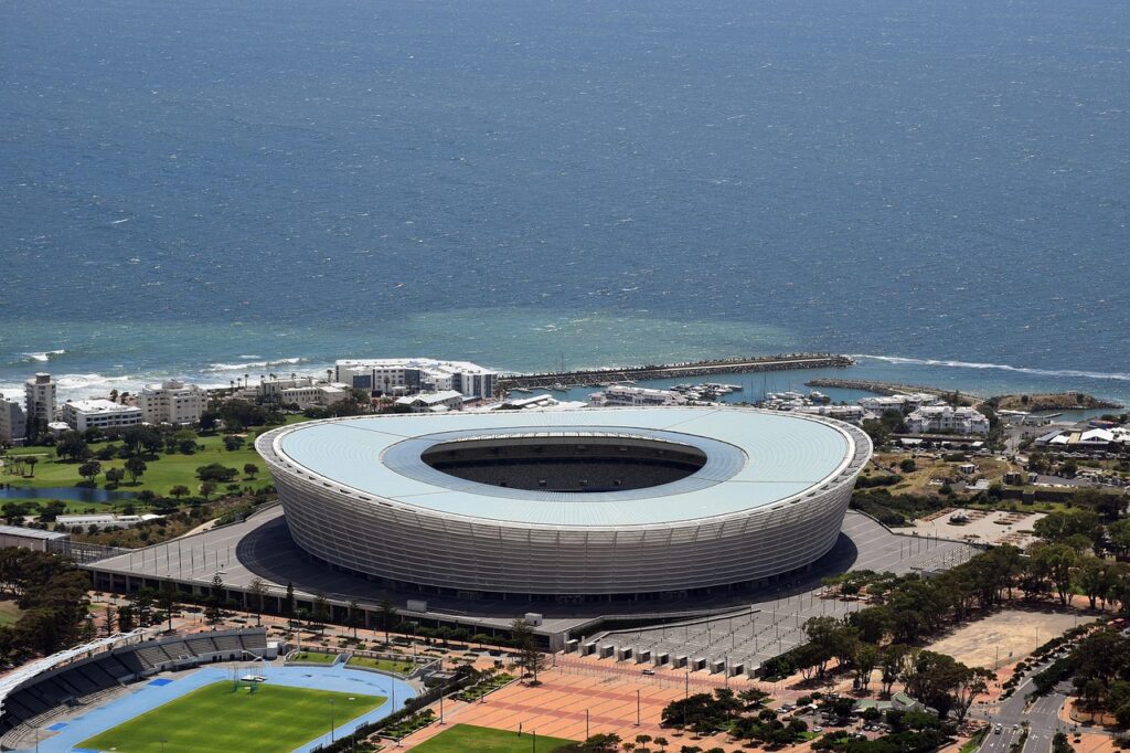 Esta imagem aérea apresenta o icônico Estádio Cape Town (Green Point Stadium) em destaque, com sua arquitetura circular moderna, posicionado próximo à costa do Oceano Atlântico na vibrante Cidade do Cabo, África do Sul.