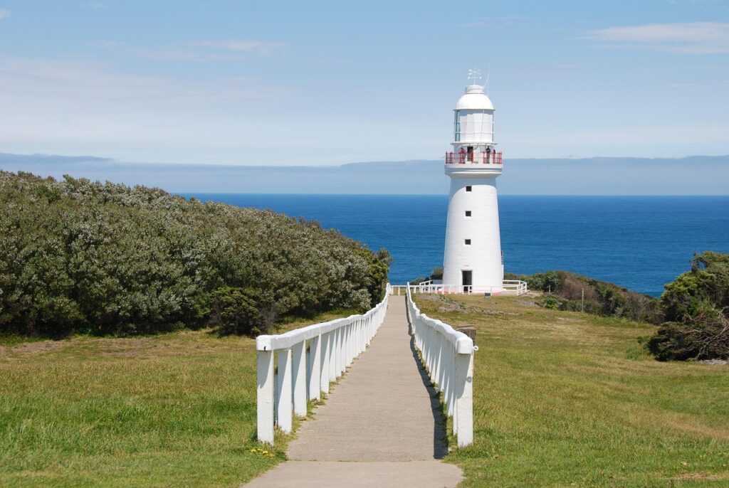 Um farol branco com detalhes vermelhos em uma colina gramada, com vista para o oceano azul e o horizonte distante.