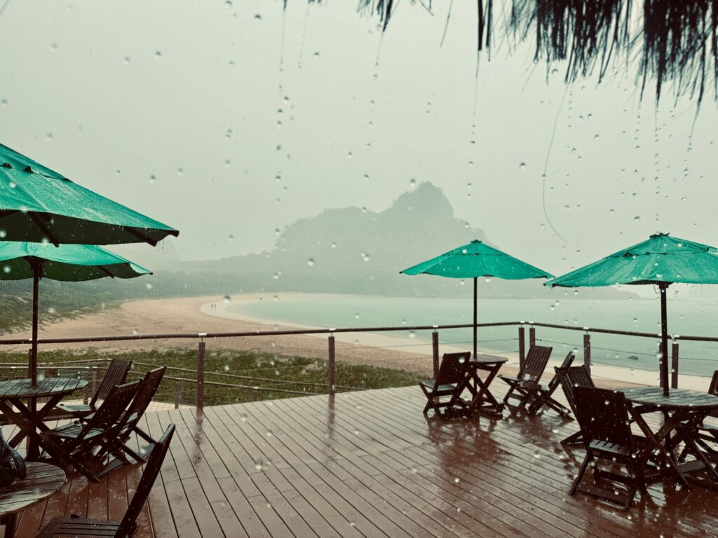 Vista de uma varanda de madeira com mesas e cadeiras de madeira e guarda-sóis verdes, com gotas de chuva visíveis no vidro, olhando para uma praia deserta com areia clara e mar calmo, e uma formação montanhosa ao fundo encoberta pela neblina ou chuva, em Fernando de Noronha.