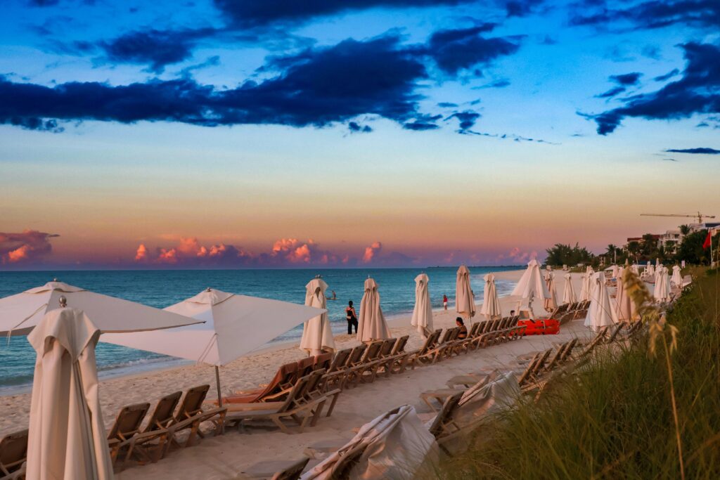 A imagem captura o entardecer na praia de Grace Bay, em Turks and Caicos, com céu multicolorido, areia branca, águas cristalinas e várias espreguiçadeiras alinhadas — cenário perfeito para quem busca um destino de luxo e tranquilidade no Caribe. Palavras-chave: Grace Bay, Turks and Caicos, praia de areia branca, pôr do sol no Caribe, viagem de luxo.