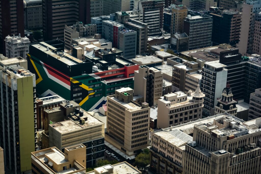 A fotografia aérea mostra a densidade urbana de Joanesburgo, África do Sul, com um destaque marcante para um edifício exibindo um grande mural da bandeira sul-africana, refletindo a essência vibrante da cidade.
