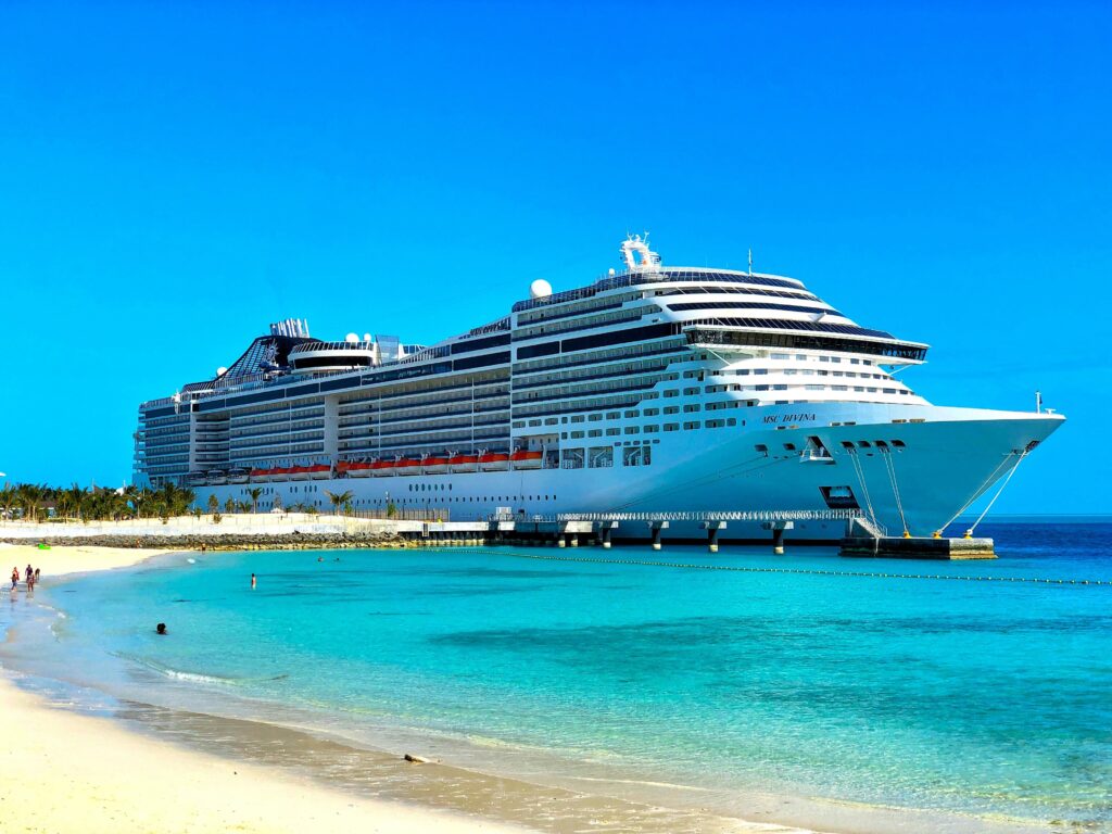 Grande navio de cruzeiro MSC Divina branco atracado ao lado de uma praia de areia clara com águas cristalinas em tons de turquesa e azul sob um céu azul sem nuvens. Pessoas estão visíveis na praia e na água.