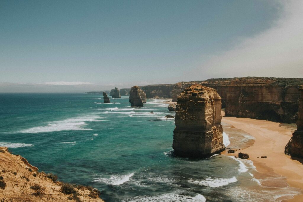 Vista panorâmica de várias pilhas de calcário ("Os Doze Apóstolos") em águas oceânicas azul-turquesa, com ondas quebrando na base. À direita, falésias costeiras íngremes e uma faixa de praia arenosa são visíveis. O céu é claro, com uma névoa leve no horizonte.
