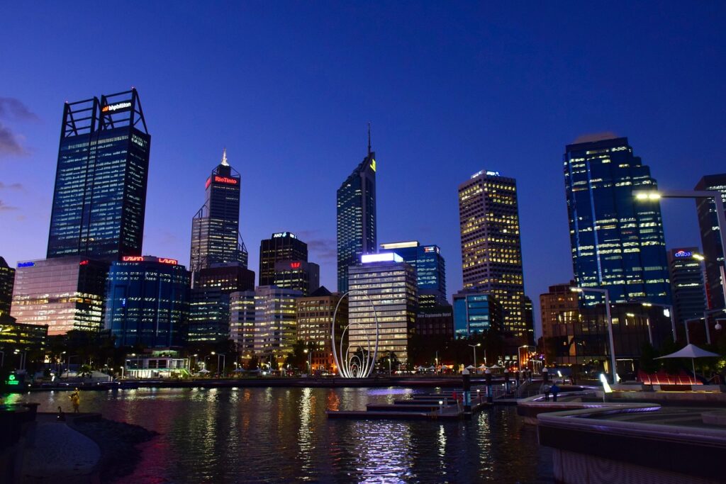 Vista noturna do horizonte da cidade de Perth, com múltiplos arranha-céus iluminados em diferentes tons de azul, verde e amarelo. As luzes dos edifícios se refletem na superfície escura de um corpo d'água em primeiro plano, onde algumas plataformas e embarcações são visíveis. O céu é de um azul profundo, indicando o anoitecer.