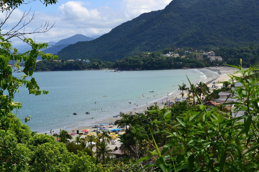 Vista panorâmica aérea da Praia Grande em Ubatuba, São Paulo, mostrando a ampla faixa de areia lotada de guarda-sóis coloridos e pessoas, com o mar calmo e barcos na baía, ladeada por montanhas cobertas de mata atlântica. A imagem destaca a infraestrutura e a popularidade da praia, ideal para SEO sobre turismo em Ubatuba e praias movimentadas.