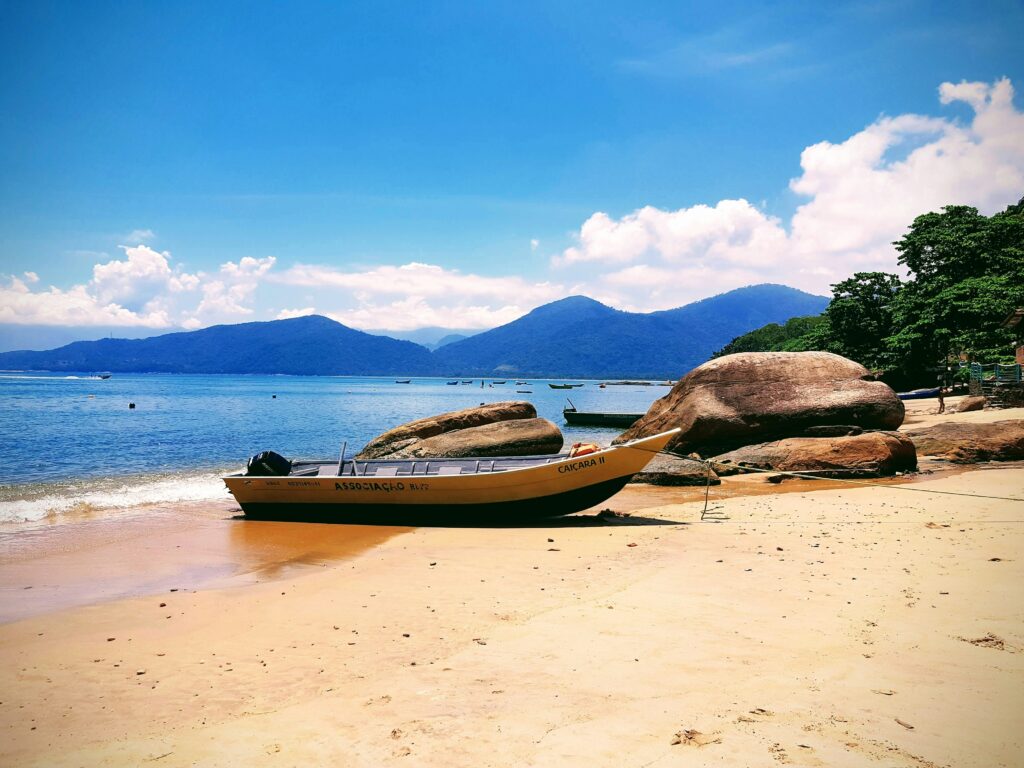 Praia de Picinguaba em Ubatuba, litoral norte de São Paulo, exibindo uma pequena embarcação de pesca na areia clara, grandes rochas arredondadas na orla, e montanhas cobertas por Mata Atlântica ao fundo sob um céu azul com poucas nuvens. Ideal para ilustrar a beleza natural e a atmosfera serena das praias de Ubatuba.