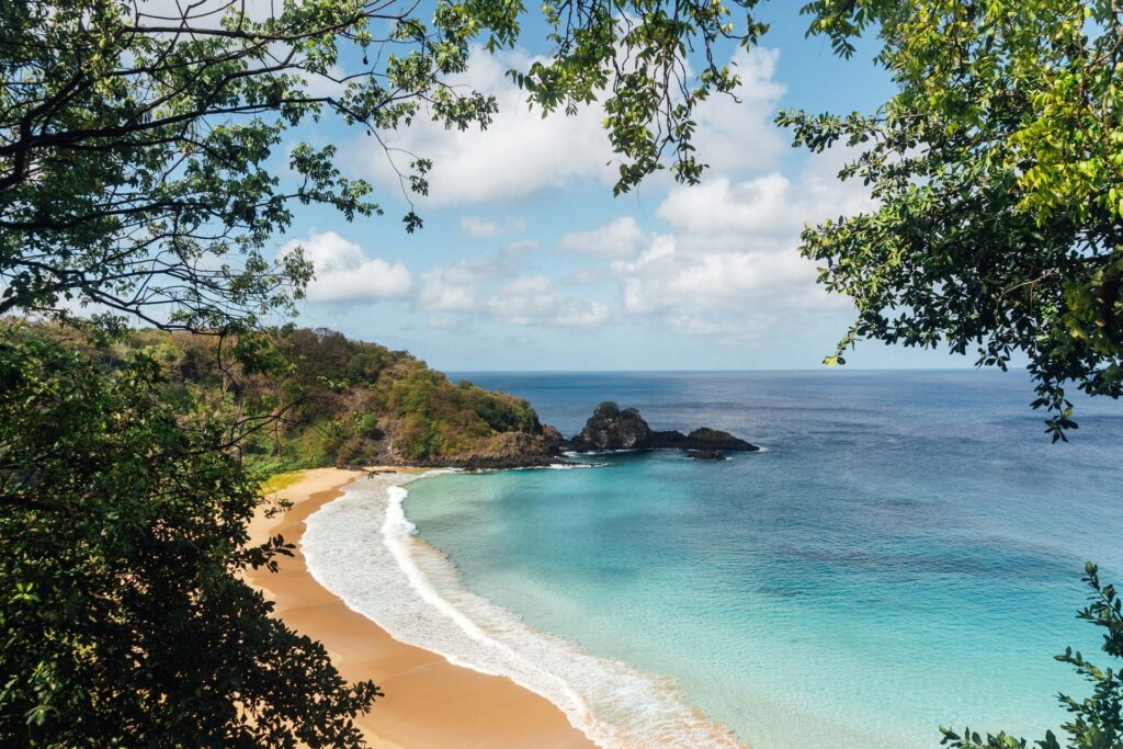 Vista aérea da Praia do Sancho em Fernando de Noronha, Brasil, mostrando a faixa de areia clara, o mar azul-turquesa com ondas suaves e a vegetação exuberante da ilha emoldurando a paisagem sob um céu parcialmente nublado.