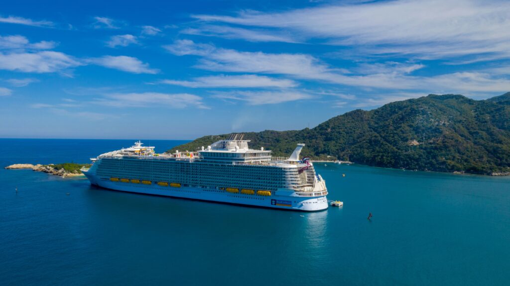 Vista aérea de um enorme navio de cruzeiro branco da Royal Caribbean, o Oasis of the Seas, ancorado em uma baía azul clara. Ao fundo, montanhas verdes e uma pequena ilha com rochas. O céu é azul com nuvens esparsas.
