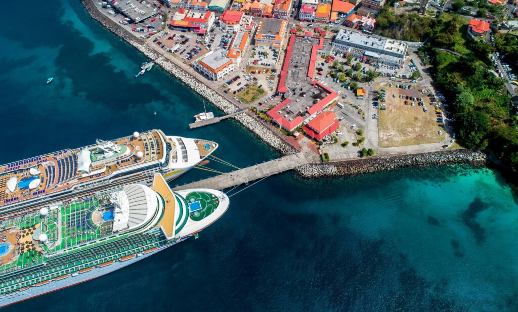 Imagem aérea do Melville Street Cruise Terminal, em St. George’s (Grenada), com dois navios de cruzeiro ancorados ao lado do centro histórico de telhados vermelhos—parada chave para roteiros pelo Caribe graças ao fácil acesso a praias, compras duty-free e excursões pela ilha. Palavras-chave: cruzeiro no Caribe, porto de Grenada, St. George’s cruise terminal, parada de navios, destino caribenho.