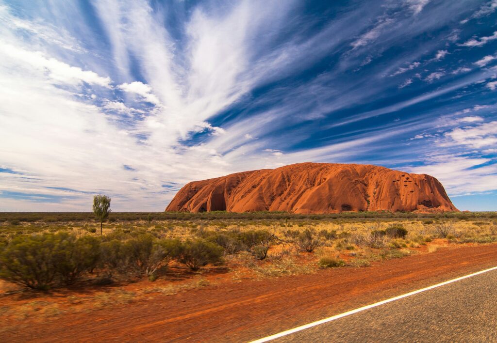 Fotografia panorâmica de Uluru (Ayers Rock), uma grande formação rochosa de arenito avermelhado, em paisagem árida com vegetação rasteira e estrada de terra em primeiro plano, sob céu azul com nuvens brancas.