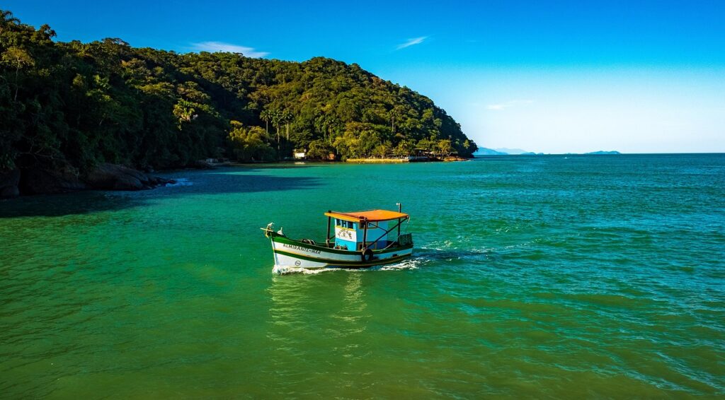 Barco de pesca tradicional com toldo laranja flutuando em águas verde-esmeralda em Ubatuba, com floresta tropical densa e montanhas ao fundo sob um céu azul claro. Ideal para representar a tranquilidade e a natureza exuberante das praias de Ubatuba, São Paulo.