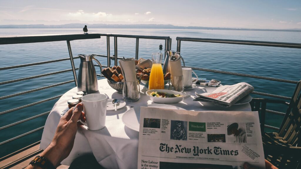 Cena de café da manhã ao ar livre em um convés de navio ou barco. Uma pessoa segura uma xícara de café enquanto um jornal The New York Times está aberto sobre a mesa branca, com suco, pães e frutas, e o oceano ao fundo.