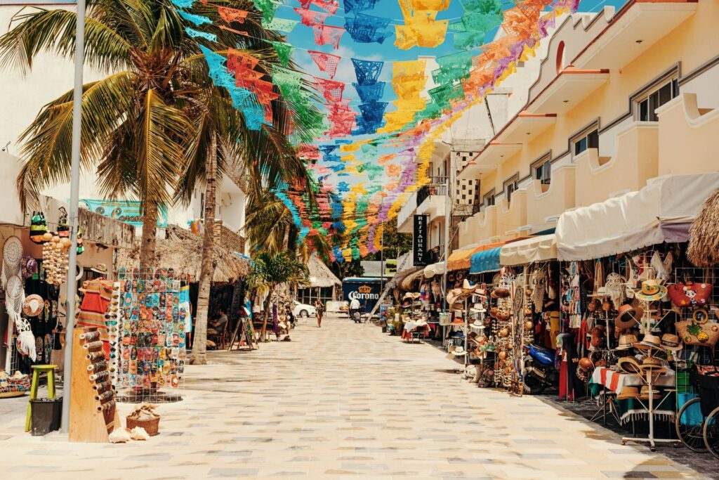 A foto retrata uma rua comercial em Cancún, México, onde barracas e lojas vendem artesanato local, lembranças e roupas sob bandeirinhas de papel picado multicoloridas. Cenário típico para compras de souvenirs no Caribe mexicano. Palavras-chave: mercado de artesanato Cancún, compras de souvenirs, rua turística mexicana, papel picado colorido, viagem ao Caribe.