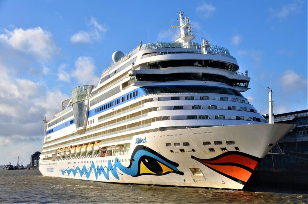 Vista frontal detalhada de um grande navio de cruzeiro branco da AIDA Cruises, o AIDAmar, atracado em um porto. A proa do navio é decorada com um grande rosto estilizado em cores vibrantes. O céu é azul com algumas nuvens.
