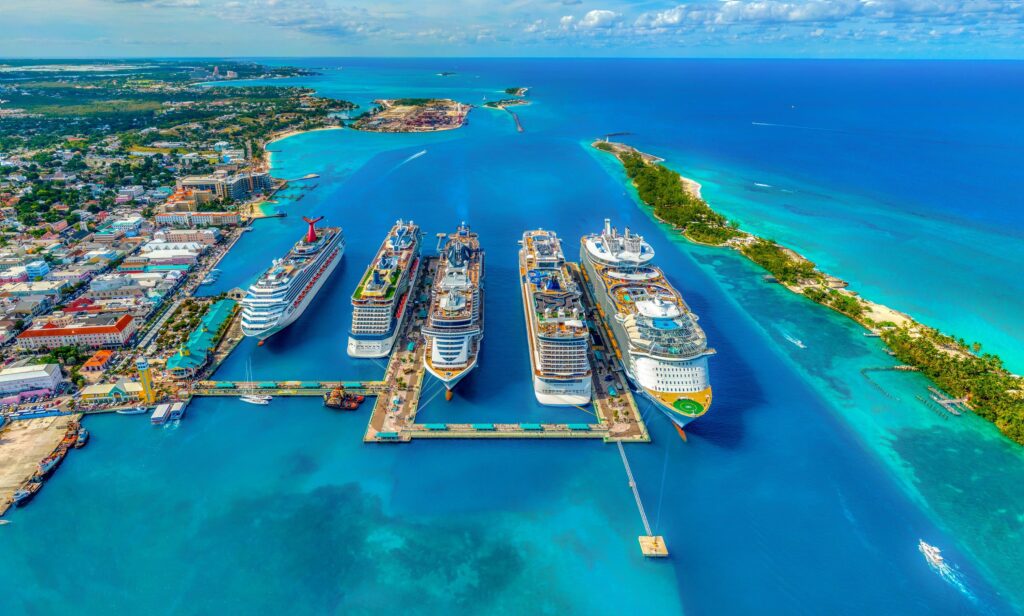 Vista aérea vibrante do porto de Nassau, nas Bahamas, mostrando seis navios de cruzeiro lado a lado em águas azul-turquesa. A imagem reforça conteúdos sobre “cruzeiros 2026”, novas rotas caribenhas e dicas de embarque em mega-navios que incluem Bahamas no itinerário.