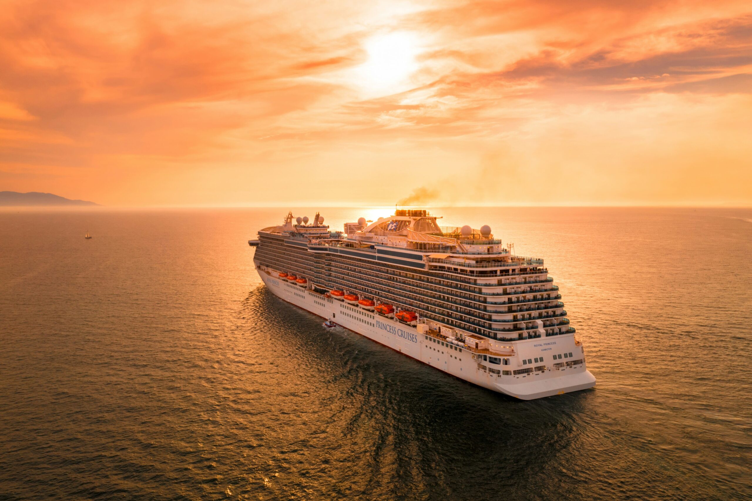 Navio de cruzeiro grande da Princess Cruises no mar aberto sob um pôr do sol laranja. A imagem mostra a popa do navio com muitos decks e botes salva-vidas.
