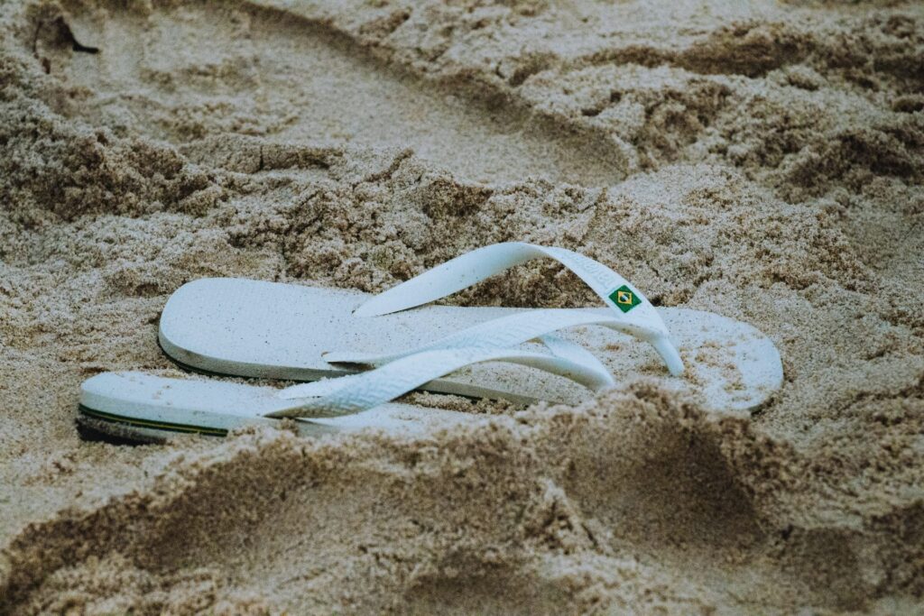 Par de chinelos brancos com detalhe da bandeira do Brasil na tira, parcialmente cobertos pela areia dourada de uma praia. A imagem evoca a sensação de férias, lazer e a cultura praiana brasileira, sendo ideal para ilustrar artigos sobre o que levar ou a atmosfera descontraída das praias de Ubatuba.