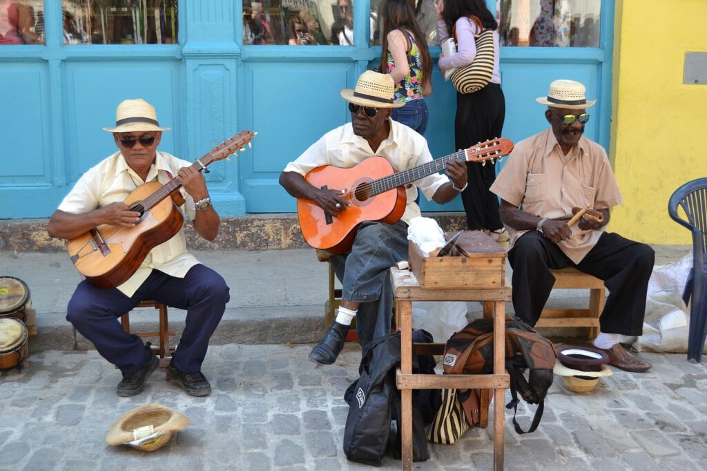 imagem mostra três músicos de rua em Havana Velha, Cuba, tocando violões e maracas em frente a prédios azuis e amarelos — cena autêntica que torna a música cubana parte essencial do turismo cultural no Caribe. Palavras-chave: Havana Vieja, música cubana ao vivo, músicos de rua, cultura em Cuba, viagem ao Caribe.