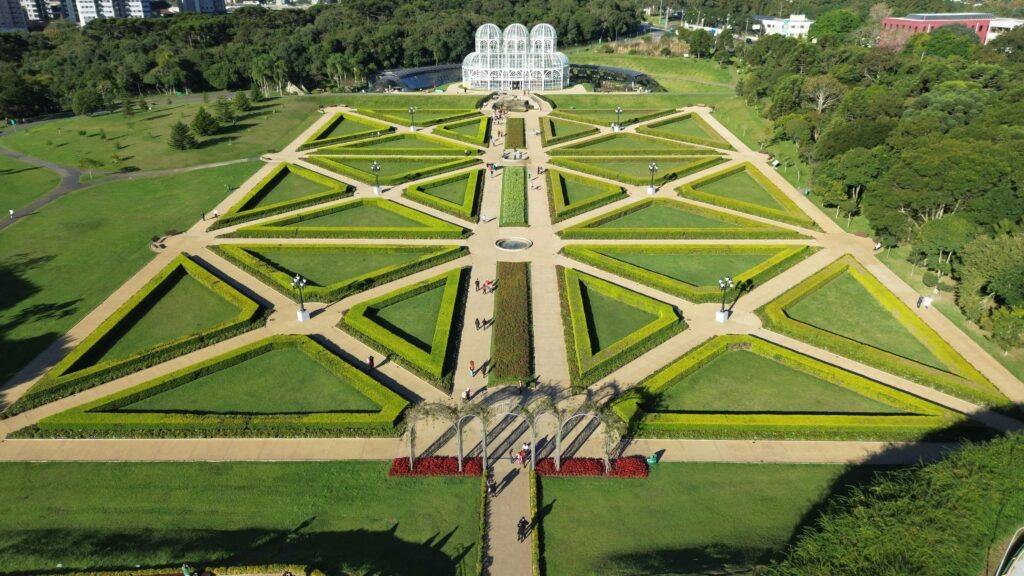 Em um ângulo superior, a imagem revela a organização paisagística do Jardim Botânico de Curitiba. Canteiros verdes, dispostos em padrões geométricos complexos, formam um tapete visual atraente. Caminhos claros cortam o jardim, guiando os visitantes. Ao fundo, a imponente estufa de vidro, com sua arquitetura delicada, destaca-se como um ponto focal. A vegetação densa e árvores ao redor emolduram o jardim, criando uma atmosfera de tranquilidade e beleza natural, imperdível para quem visita Curitiba.