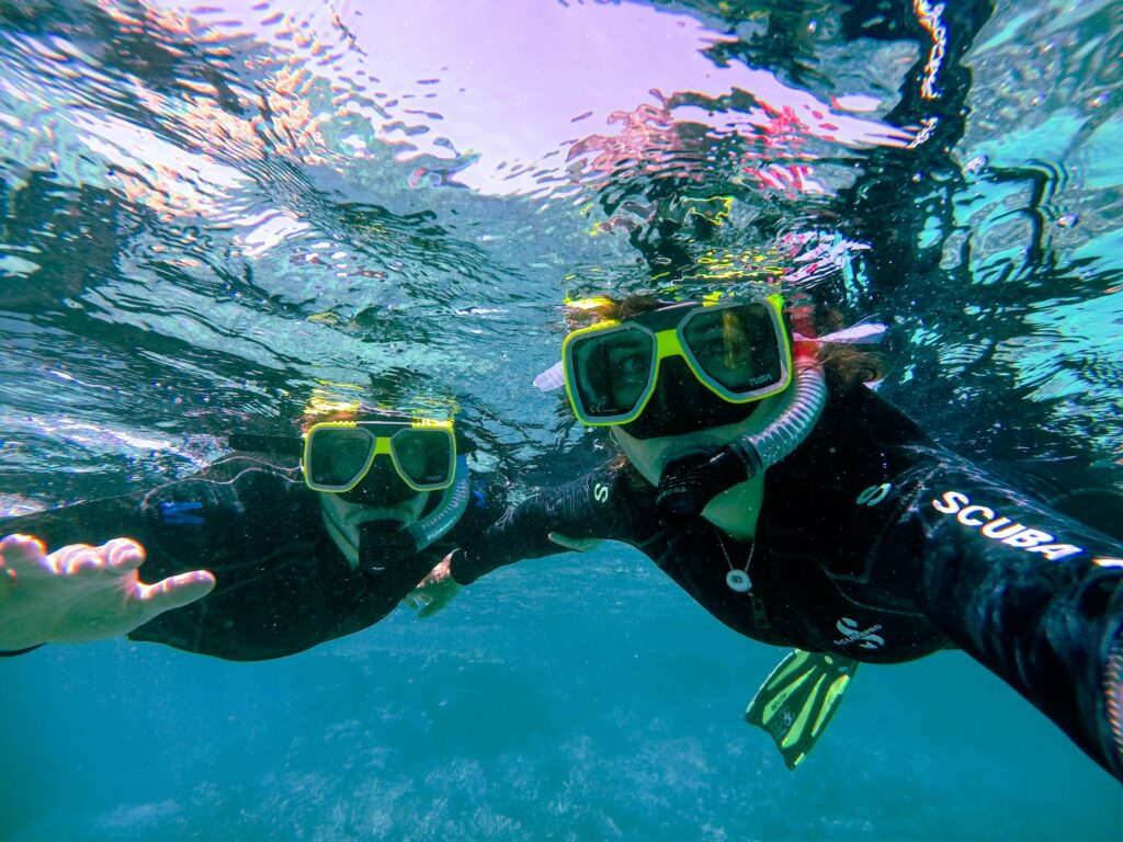 A foto mostra dois viajantes praticando snorkeling no Mar do Caribe, equipados com máscaras e trajes de neoprene enquanto exploram recifes e vida marinha em águas cristalinas—experiência imperdível para quem busca mergulho e turismo de aventura no Caribe. Palavras-chave: mergulho no Caribe, snorkeling, recife de coral, águas cristalinas, turismo de aventura.