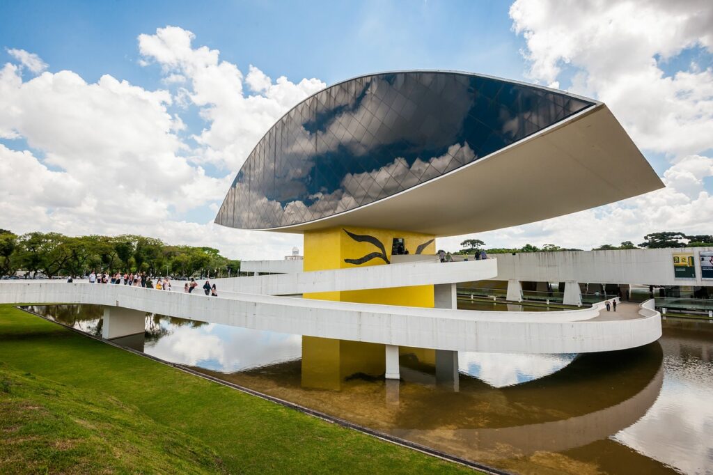 Museu Oscar Niemeyer (MON) em Curitiba, Paraná, Brasil, com sua estrutura principal em formato de olho e passarela sinuosa sobre a água, sob um céu azul com nuvens brancas. O edifício amarelo vibrante e a fachada espelhada refletem a paisagem ao redor, destacando a arquitetura moderna de Oscar Niemeyer. Pessoas são vistas caminhando na passarela e nas áreas verdes adjacentes ao museu, indicando um local turístico e cultural movimentado.