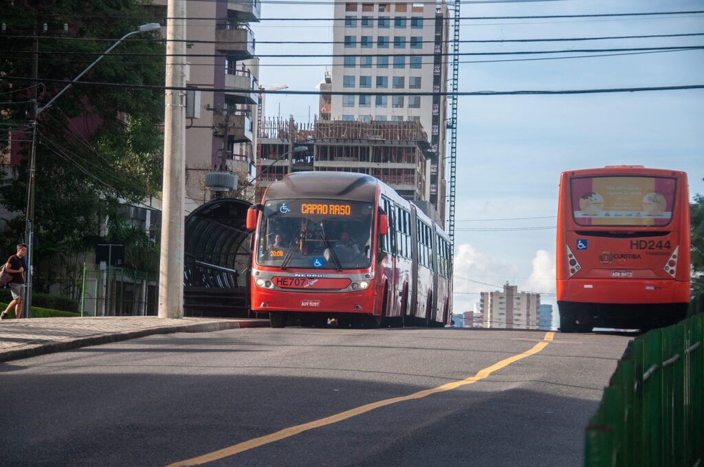 Dois ônibus vermelhos do sistema BRT de Curitiba, Brasil, em uma rua urbana. O ônibus em primeiro plano, com destino "CAPÃO RASO", se aproxima, enquanto a traseira de outro ônibus é visível à direita. Edifícios altos e estruturas urbanas compõem o fundo da imagem, destacando a infraestrutura de transporte público da capital paranaense. Um pedestre caminha na calçada à esquerda, e um poste de iluminação é visível, complementando a cena de um dia claro na cidade.