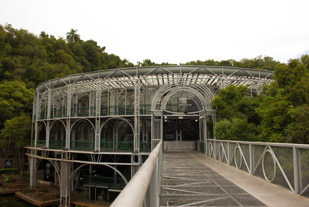 A Ópera de Arame em Curitiba, Paraná, Brasil, um teatro de estrutura tubular metálica e teto transparente, cercado por uma densa floresta verde. Uma passarela de acesso em metal leva até a entrada principal do edifício, que se integra harmoniosamente com a paisagem natural. A arquitetura única do local destaca-se como um ponto turístico e cultural, refletindo a luz do dia através de sua estrutura vazada.