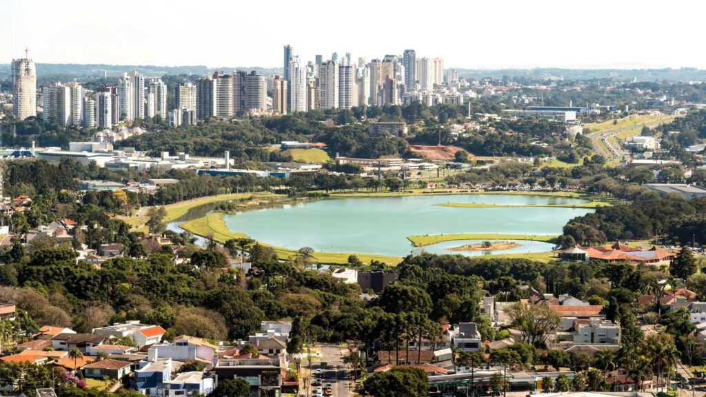 Vista aérea do Parque Barigui em Curitiba, destacando o grande lago cercado por vegetação e, ao fundo, o denso horizonte de edifícios do centro da cidade sob um céu claro.