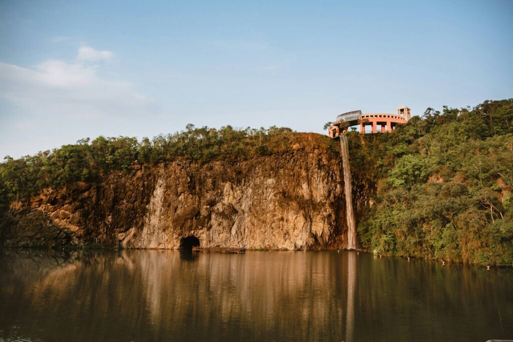 Vista panorâmica do Parque Tanguá em Curitiba, mostrando a cachoeira artificial caindo do paredão de rocha, o lago na base e o edifício do mirante no topo da colina sob um céu azul.