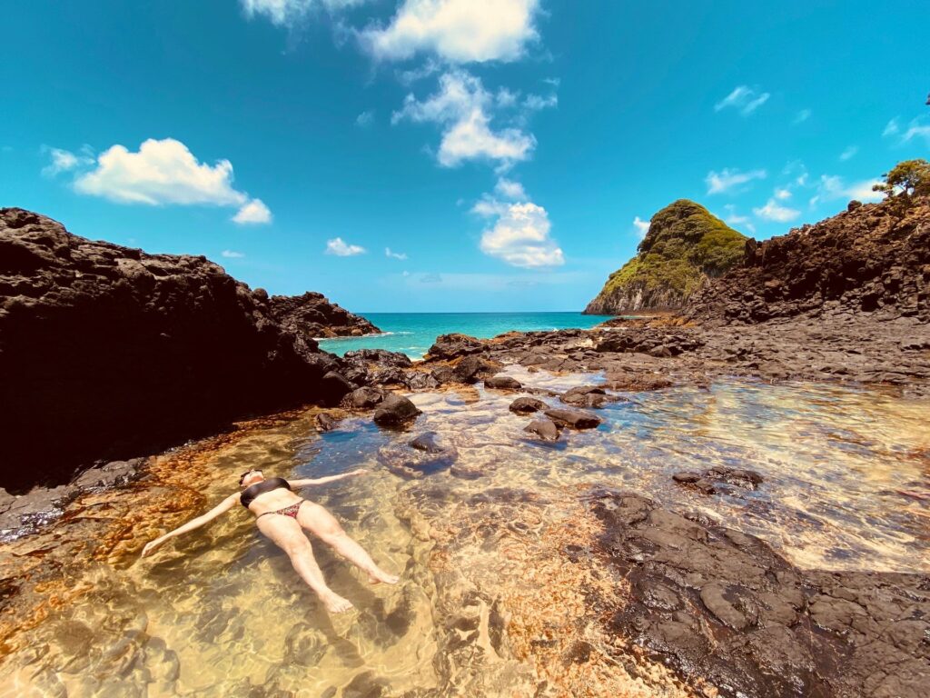Uma pessoa deitada de costas flutuando em uma piscina natural de águas claras, cercada por formações rochosas escuras, com o mar azul-turquesa e uma ilha montanhosa ao fundo sob um céu azul com nuvens brancas, em Fernando de Noronha.