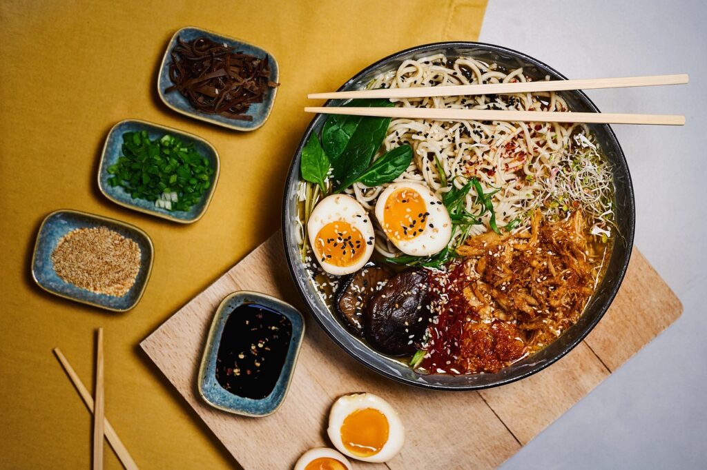 Tigela de ramen japonês com macarrão, carne, ovo cozido, cogumelos e cebolinha, acompanhado de molhos e hashi.