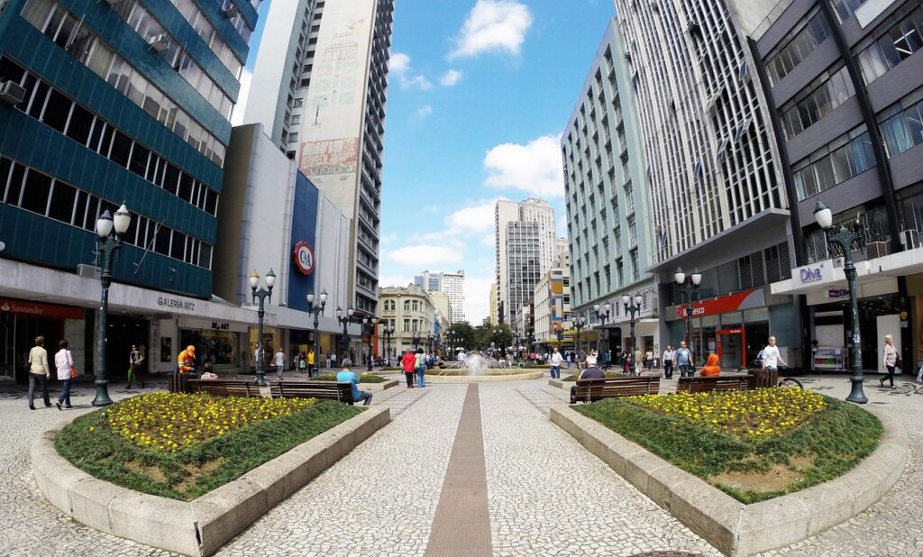 Vista panorâmica da Rua XV de Novembro em Curitiba, Paraná, uma rua de pedestres arborizada com canteiros floridos e bancos. Edifícios comerciais altos ladeiam a rua, com lojas e fachadas urbanas. Pessoas circulam pelo calçadão de pedras portuguesas, e ao fundo, uma fonte e mais prédios completam a cena urbana vibrante do centro de Curitiba, destacando a arquitetura e a vida da cidade.