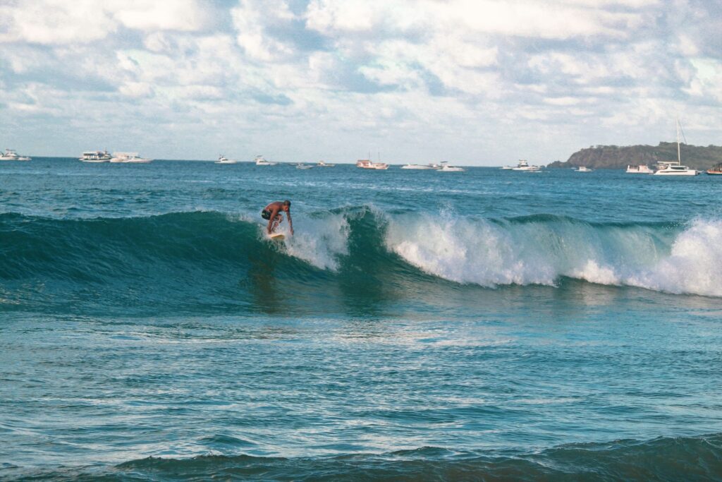Um surfista de costas sobre uma prancha, surfando uma onda quebrando em águas azuis-esverdeadas, com várias embarcações e uma faixa de terra com vegetação ao fundo sob um céu parcialmente nublado, em Fernando de Noronha, Brasil.