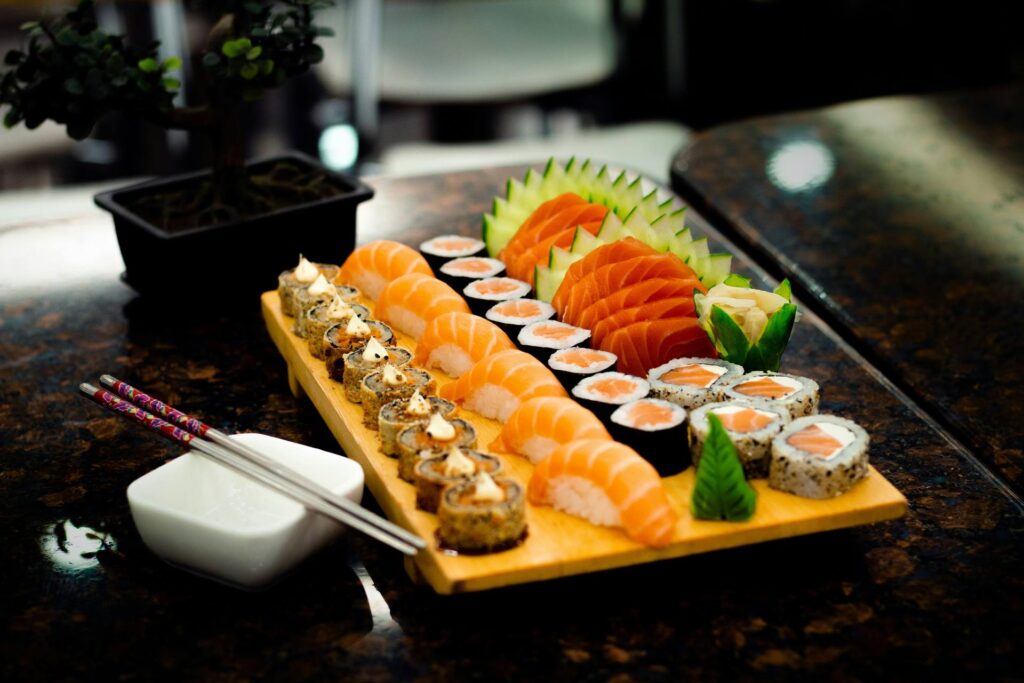 Combinado de sushi, sashimi de salmão, nigiri e rolls servidos em uma travessa de madeira, com hashi e um bonsai ao lado.