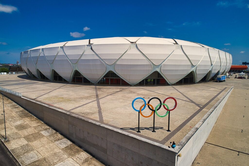 Vista exterior da Arena da Amazônia em Manaus, um grande estádio branco com arquitetura moderna, e os icônicos anéis olímpicos coloridos em primeiro plano, sob um céu azul.