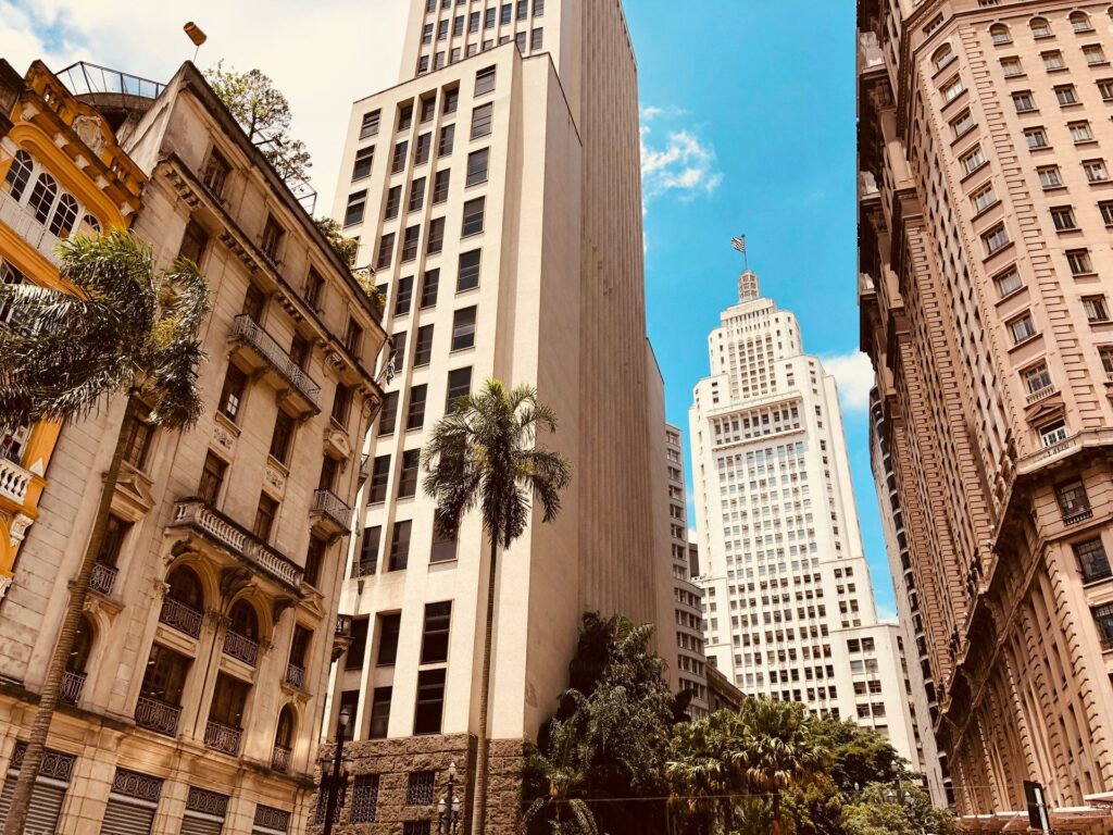 Vista de baixo para cima de edifícios históricos e modernos no centro de São Paulo, com o imponente Edifício Altino Arantes (Banespão) ao fundo, sob um céu azul com nuvens esparsas, palmeiras e detalhes arquitetônicos em prédios antigos, ideal para ilustrar a diversidade arquitetônica da capital paulista.