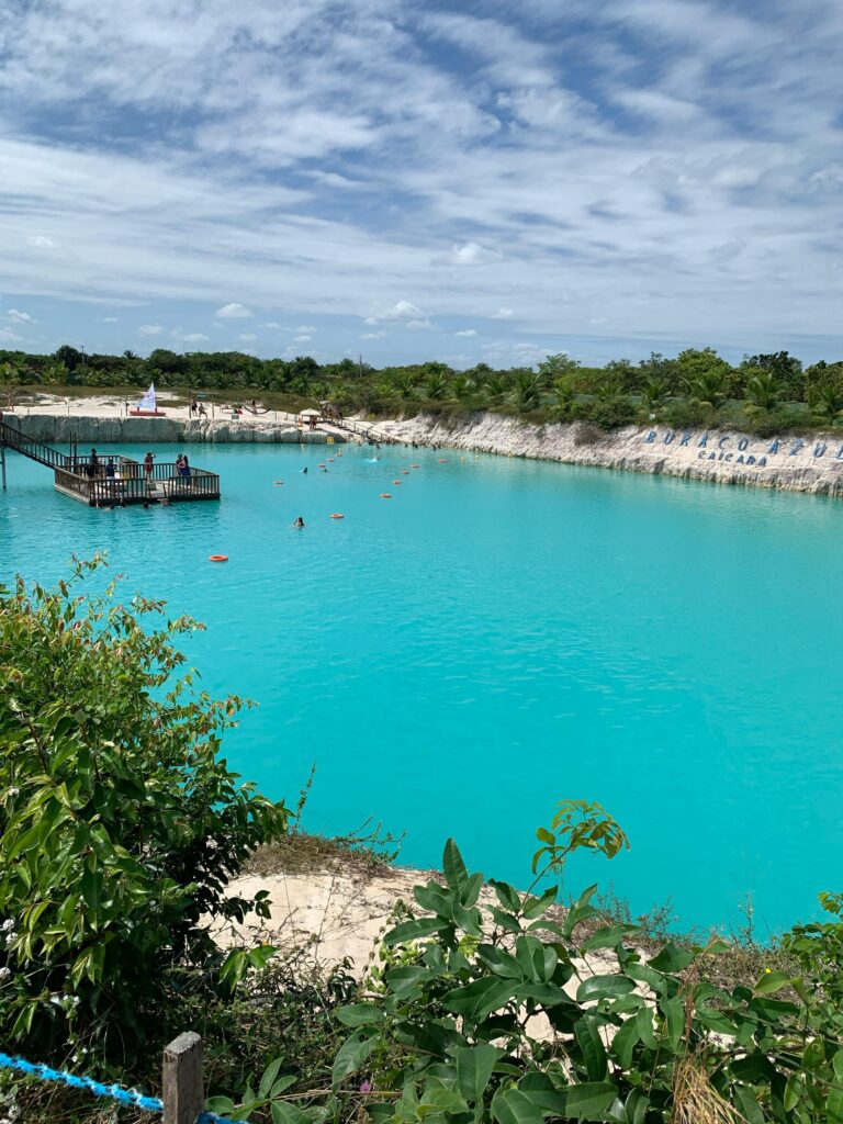 Vista panorâmica do Buraco Azul Caiçara em Jericoacoara, Ceará, uma lagoa artificial de águas azul-turquesa vibrantes, com pessoas nadando e relaxando em um deck de madeira flutuante. A paisagem é cercada por vegetação rasteira e o céu tem nuvens esparsas, com uma placa de "Buraco Azul Caiçara" visível ao fundo.