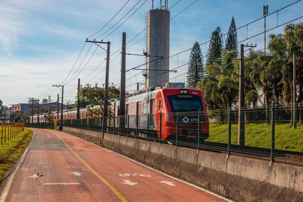 Trem moderno da CPTM (Companhia Paulista de Trens Metropolitanos) de cor vermelha e cinza em movimento sobre trilhos, com uma ciclovia de asfalto vermelho ao lado em primeiro plano e vegetação, postes de fiação e uma torre de água ao fundo, sob um céu azul, representando o transporte público e a infraestrutura de mobilidade em São Paulo.