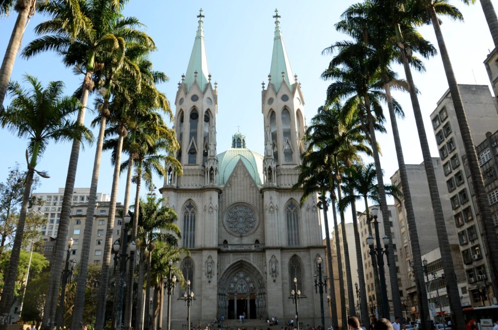 Vista frontal da Catedral Metropolitana de São Paulo (Catedral da Sé) sob um céu azul, com palmeiras altas em primeiro plano e prédios ao redor, destacando sua arquitetura gótica e grandiosidade, ideal para ilustrar artigos sobre turismo em São Paulo.