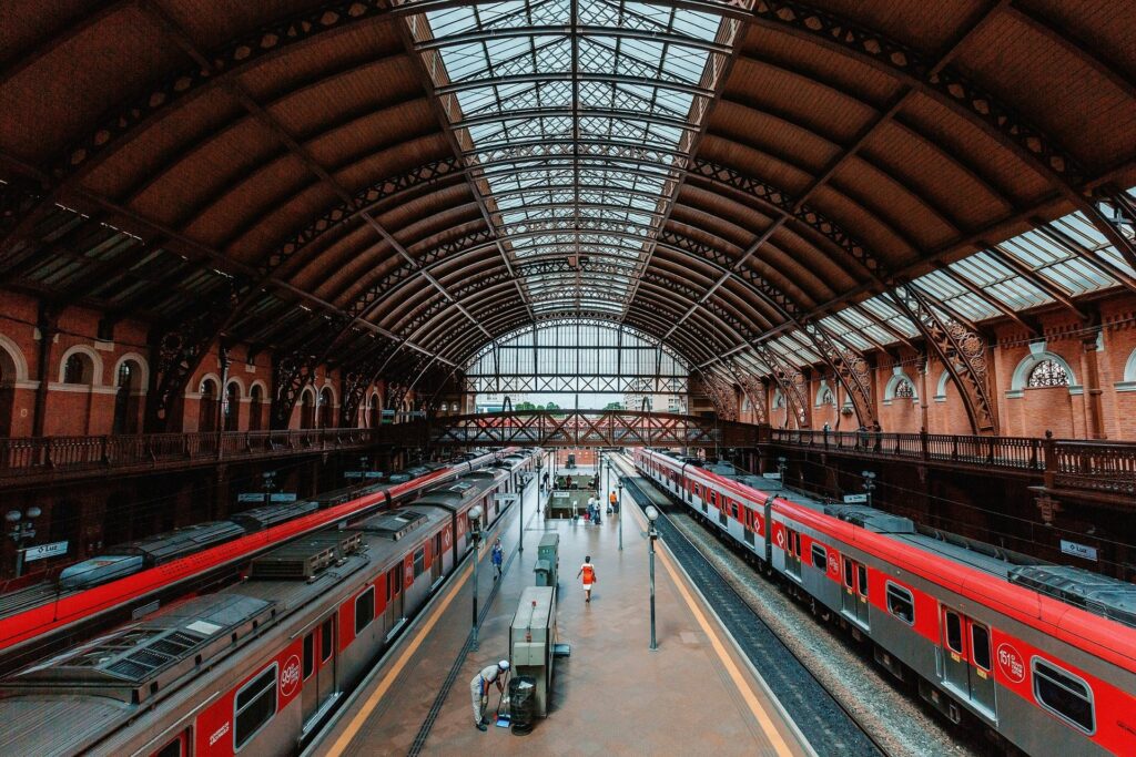 Interior grandioso da Estação da Luz em São Paulo, mostrando plataformas com trens vermelhos estacionados, sob uma impressionante cobertura metálica e de vidro que revela sua arquitetura histórica, com algumas pessoas aguardando, ideal para ilustrar artigos sobre transporte e pontos turísticos na capital paulista.