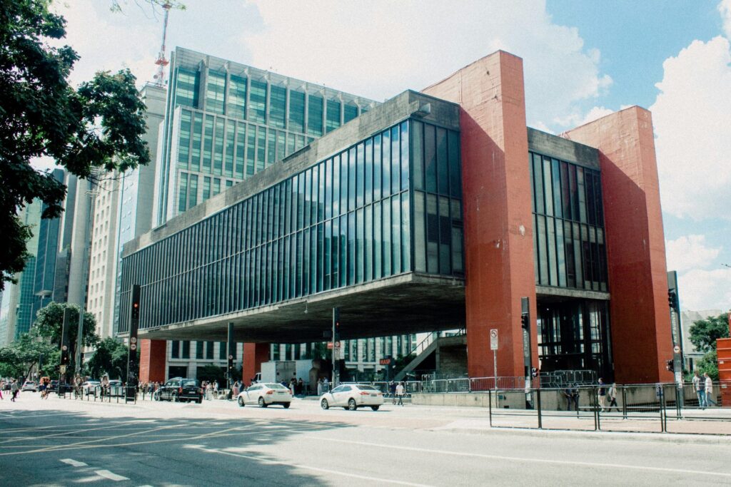 Vista externa do Museu de Arte de São Paulo (MASP) na Avenida Paulista, com sua estrutura de concreto e vidro elevada por pilares vermelhos, veículos e pedestres transitando na rua em frente, sob um céu parcialmente nublado, ideal para ilustrar roteiros culturais em São Paulo.