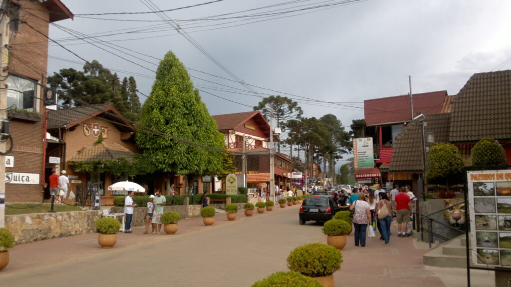 Vista da avenida em Monte Verde, Minas Gerais, com turistas e moradores caminhando entre lojas e restaurantes de arquitetura europeia, árvores altas e postes de iluminação, refletindo o ambiente acolhedor da cidade.