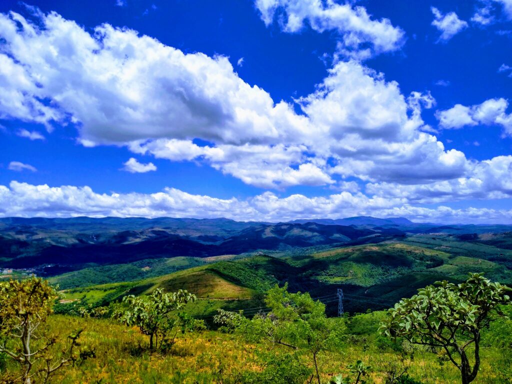 Vista panorâmica de vastas colinas e montanhas cobertas por vegetação verdejante sob um céu azul vibrante com nuvens brancas.
