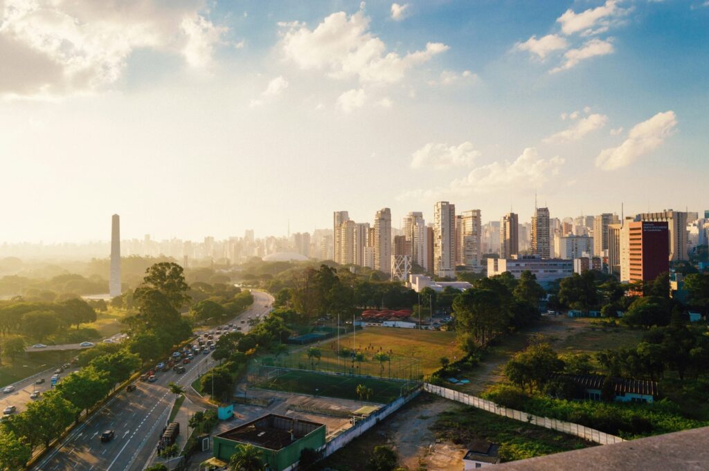 Vista aérea panorâmica de São Paulo ao entardecer, com destaque para a Avenida 23 de Maio, o Obelisco do Ibirapuera e arranha-céus, sob um céu com nuvens esparsas e luz solar, ideal para ilustrar roteiros de viagem na capital paulista.