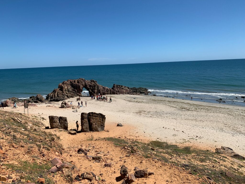 A famosa Pedra Furada em Jericoacoara, Ceará, uma formação rochosa natural com um arco no meio, situada em uma praia de areia clara, com o oceano azul ao fundo e o céu claro. Pessoas são visíveis na praia, explorando a área.
