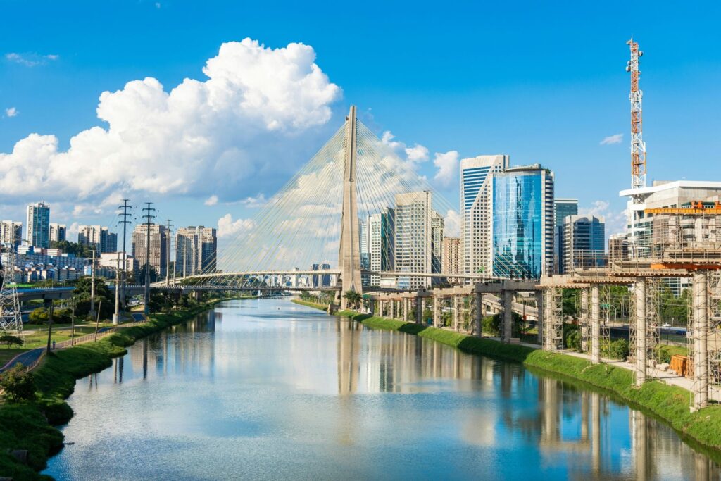 Vista panorâmica da Ponte Estaiada (Octávio Frias de Oliveira) em São Paulo, atravessando o Rio Pinheiros com seus cabos simétricos, rodeada por arranha-céus modernos sob um céu azul com nuvens brancas, simbolizando o progresso e a beleza urbana da metrópole, ideal para ilustrar roteiros em São Paulo.