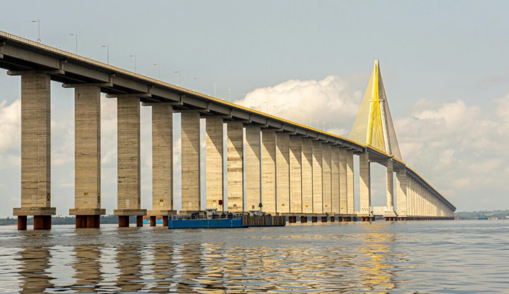 Vista lateral da Ponte Rio Negro (Ponte Jornalista Phelippe Daou) em Manaus, exibindo seus longos pilares de concreto e a torre estaiada amarela sobre as águas do rio, sob céu parcialmente nublado.